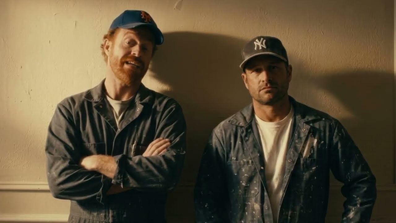 Real Pest — two exterminators in paint-spattered coveralls and ballcaps lean against a wall, one mid-pitch.